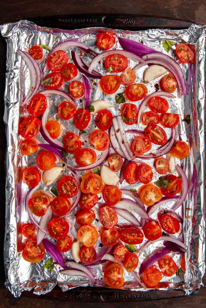 a baking sheet lined with tin foil with cherry tomatoes, red onion, and garlic on it covered in olive oil and seasoning