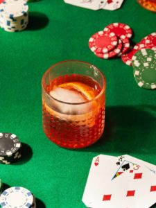 Aperol Old Fashioned cocktail in a glass tumbler on a green poker table felt with poker chips and playing cards