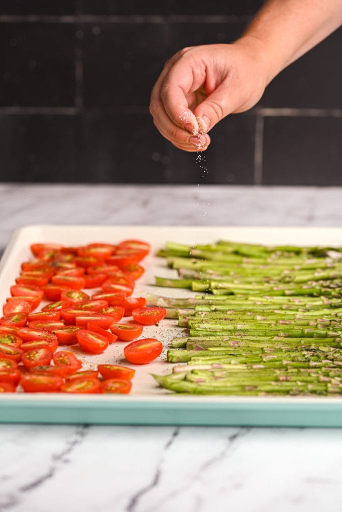 adding salt to grape tomatoes and asparagus on a sheet pan