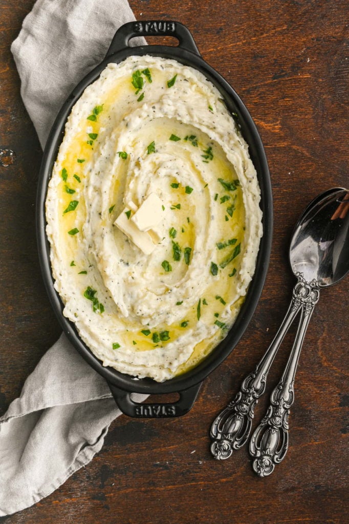 buttery mashed potatoes with herbs in a black casserole dish with two large serving spoons