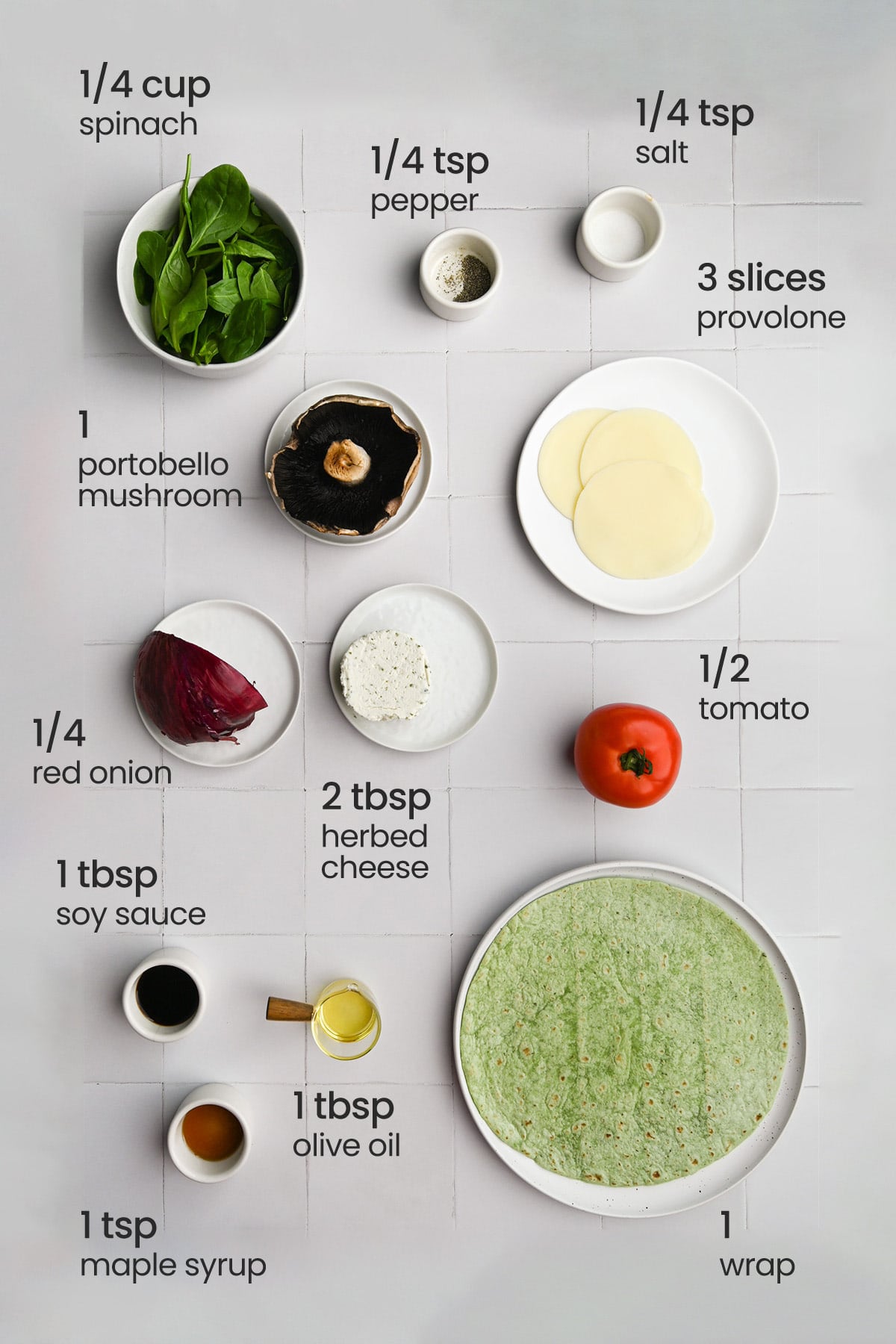Overhead view of the ingredients needed for a portobello mushroom wrap - spinach, pepper, salt, portobello mushroom, provolone cheese, red onion, herbed cheese, tomato, wrap, maple syrup, soy sauce, and olive oil.