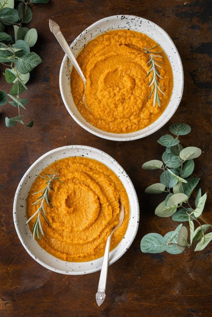 2 Bowls of Carrot Pumpkin Soup with Spoons and Surrounded by Greenery