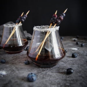 two stemless wine glasses with whiskey blues cocktail, whiskey ice, and cocktail sticks with blueberry garnish