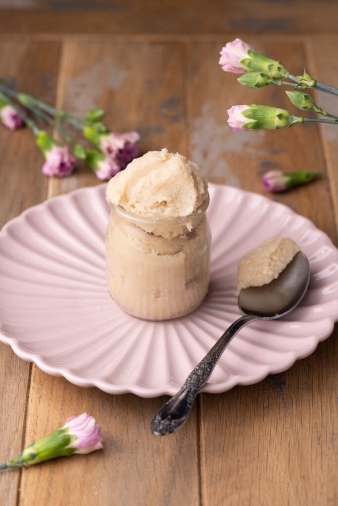 Edible sugar cookie dough in a glass jar on a pink plate with a spoonful scooped out and placed on the plate next to the jar.