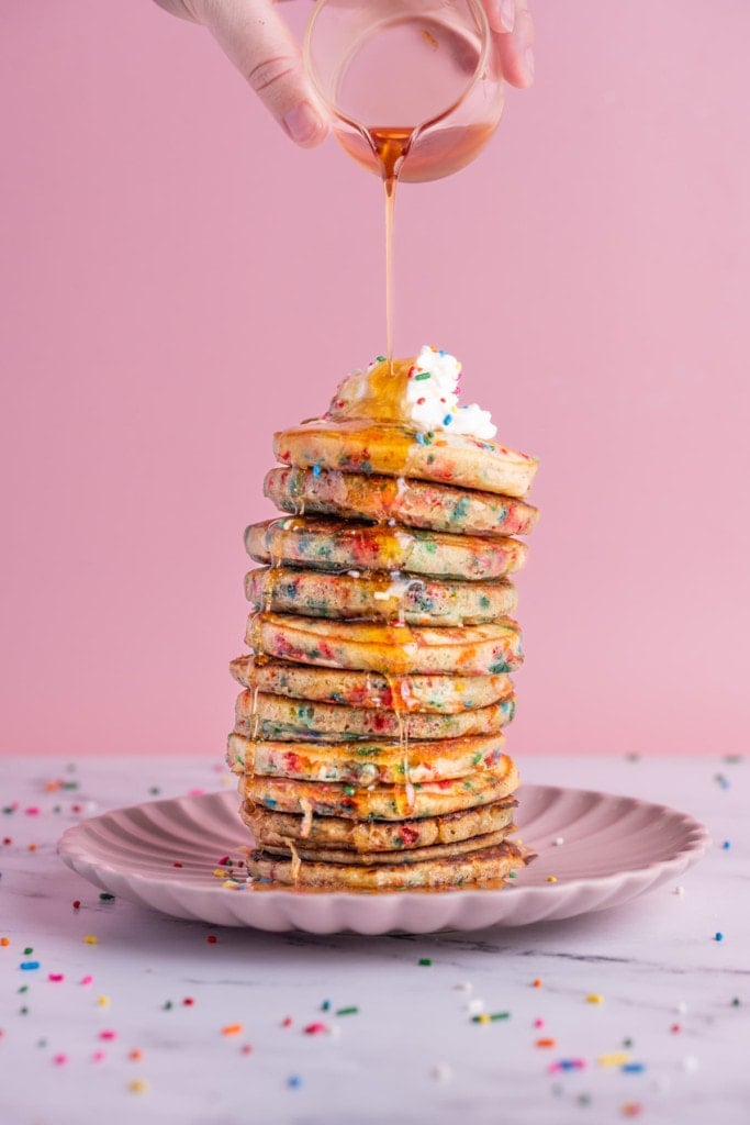 Pouring maple syrup on tall stack of Funfetti Pancakes