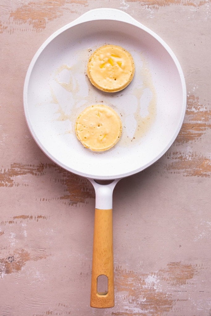 Frying two perfectly round mango pancakes in a frying pan with butter