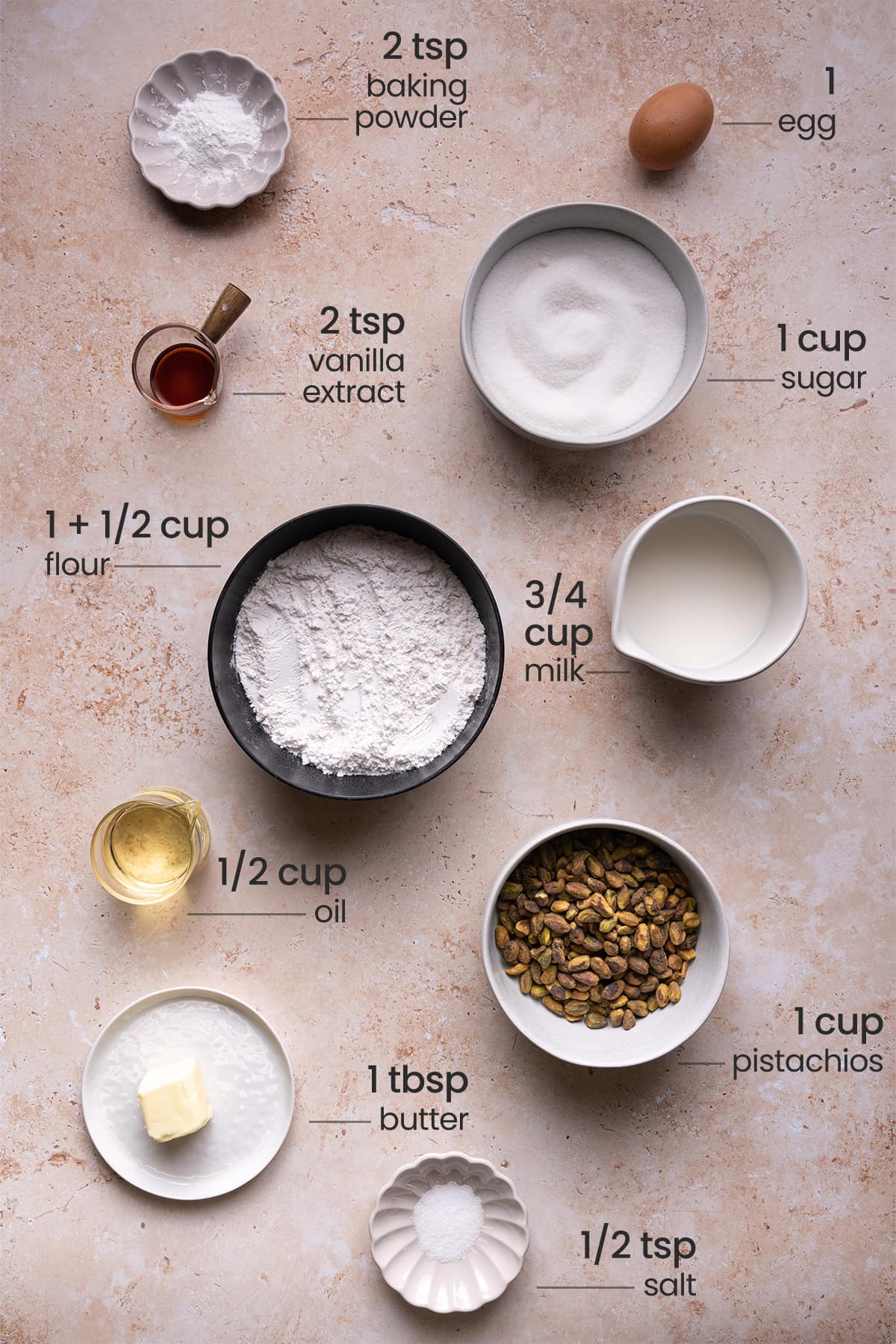 overhead view of all ingredients - baking powder, egg, vanilla extract, sugar, flour, milk, oil, pistachios, butter, salt