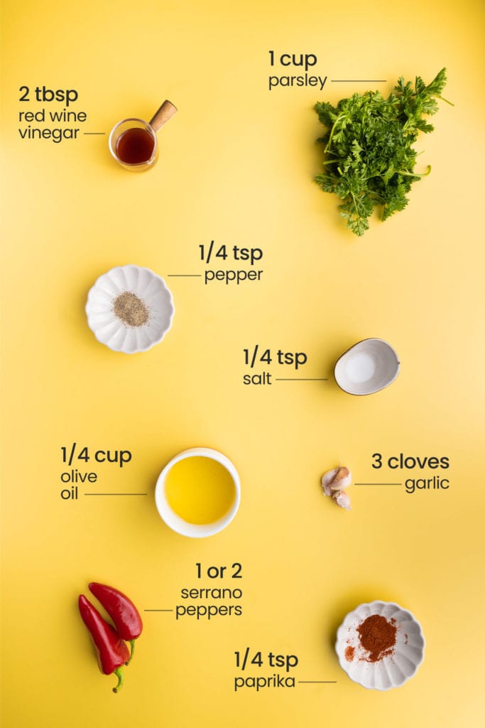 Overhead picture of all ingredients needed for Serrano Pepper Chimichurri - red wine vinegar, fresh parsley, pepper, salt, olive oil, garlic, serrano peppers, and paprika