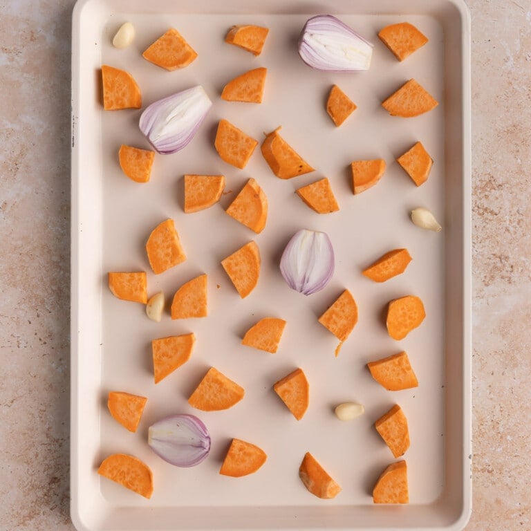 Sweet potatoes, shallots, and garlic spread out on a sheet pan