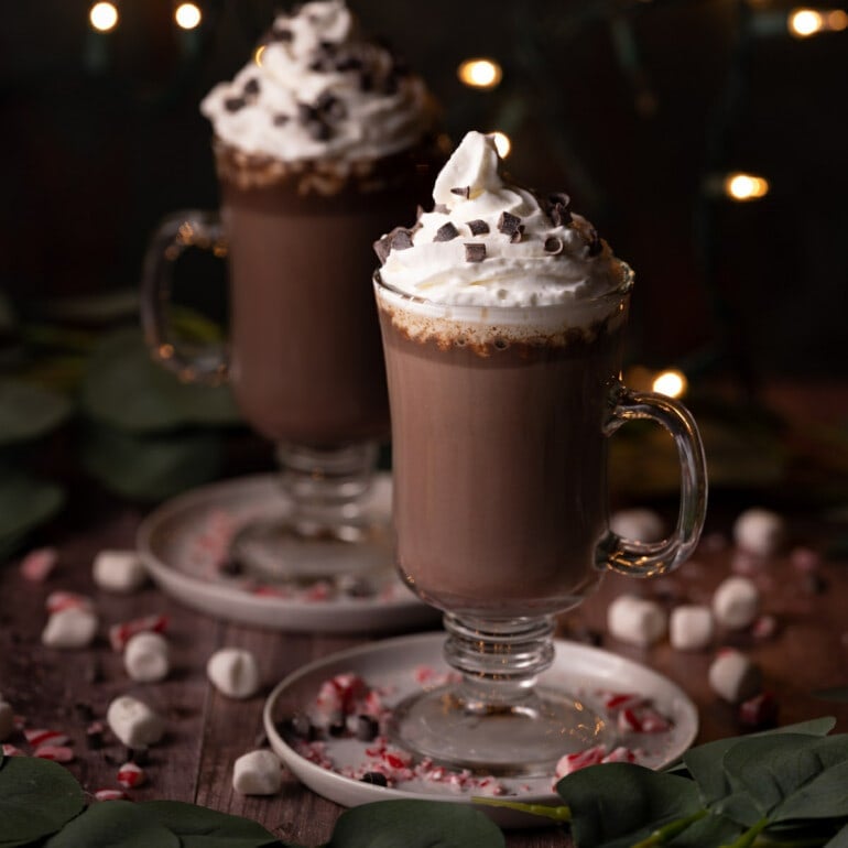 Two glass mugs of Baileys Hot Chocolate topped with Whipped Cream and chocolate curls