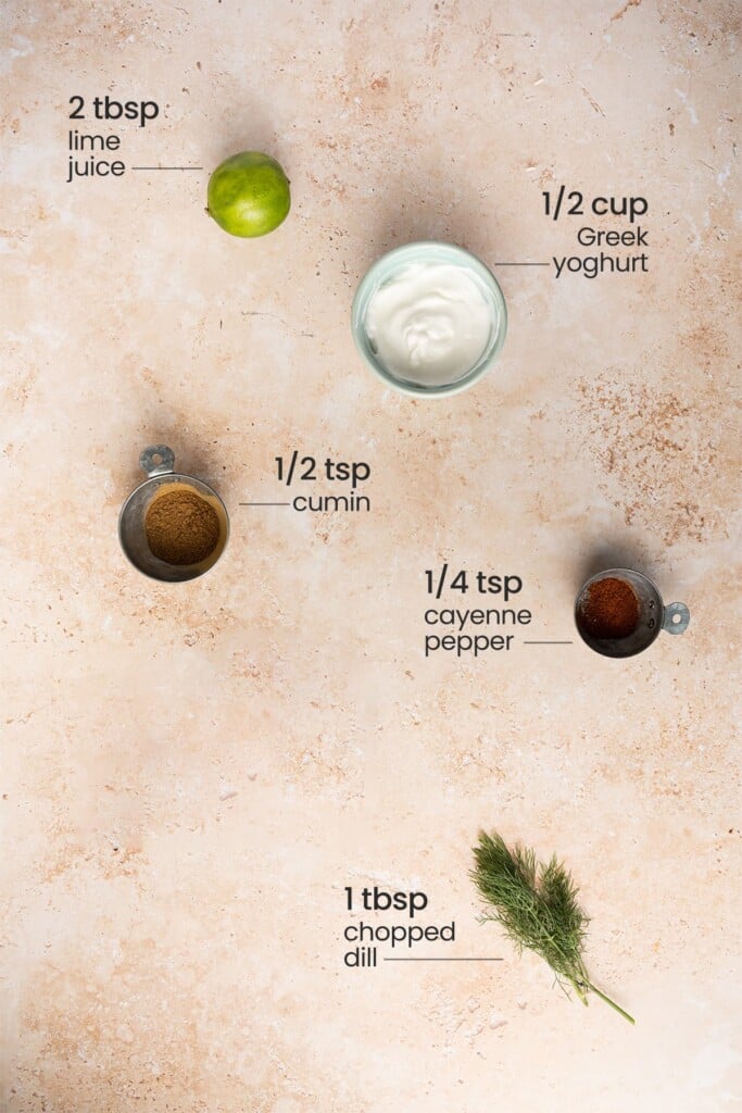 Overhead view of the ingredients needed for Greek yogurt sauce for tacos - lime, Greek yogurt, cumin, cayenne pepper, and fresh dill.
