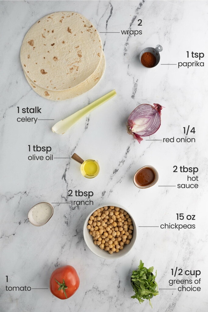 overhead view of all ingredients for buffalo chickpea wrap - wraos, paprika, celery, red onion, olive oil, hot sauce, ranch, chickpeas, tomato, greens