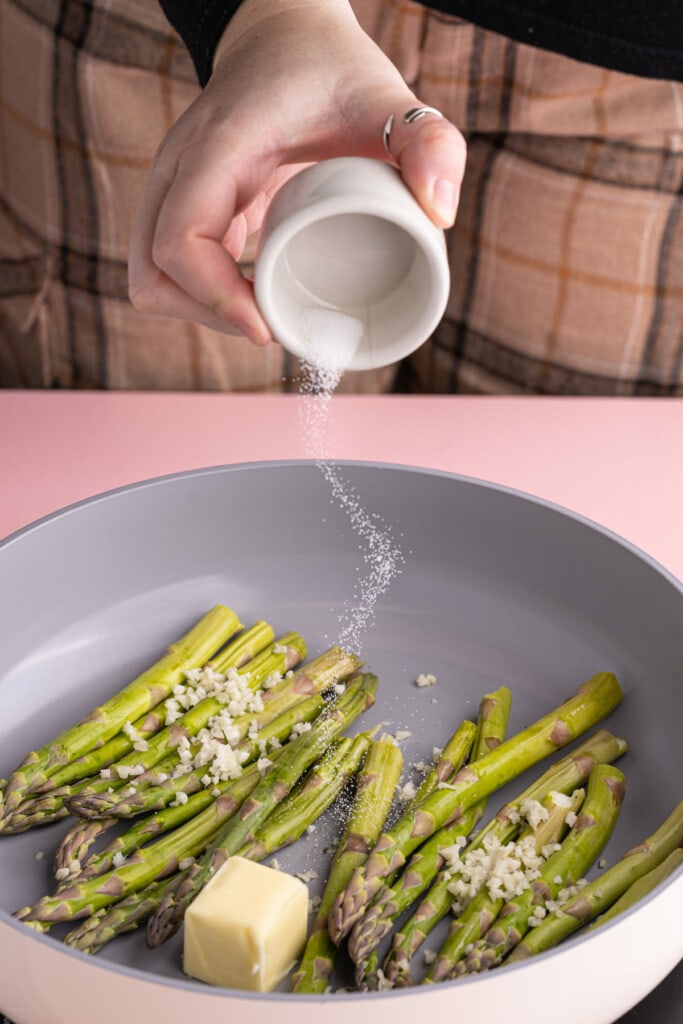 Adding salt to a pan with butter, asparagus, garlic, and pepper