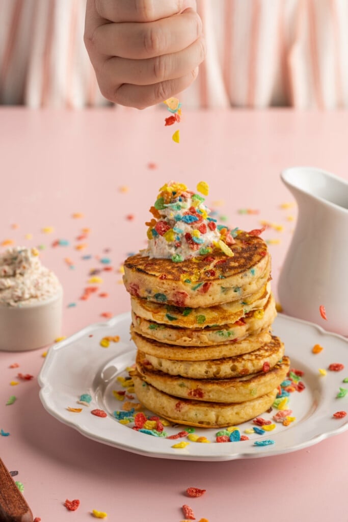 Sprinkling extra fruity pebbles on top of Fruity Pebbles Pancakes