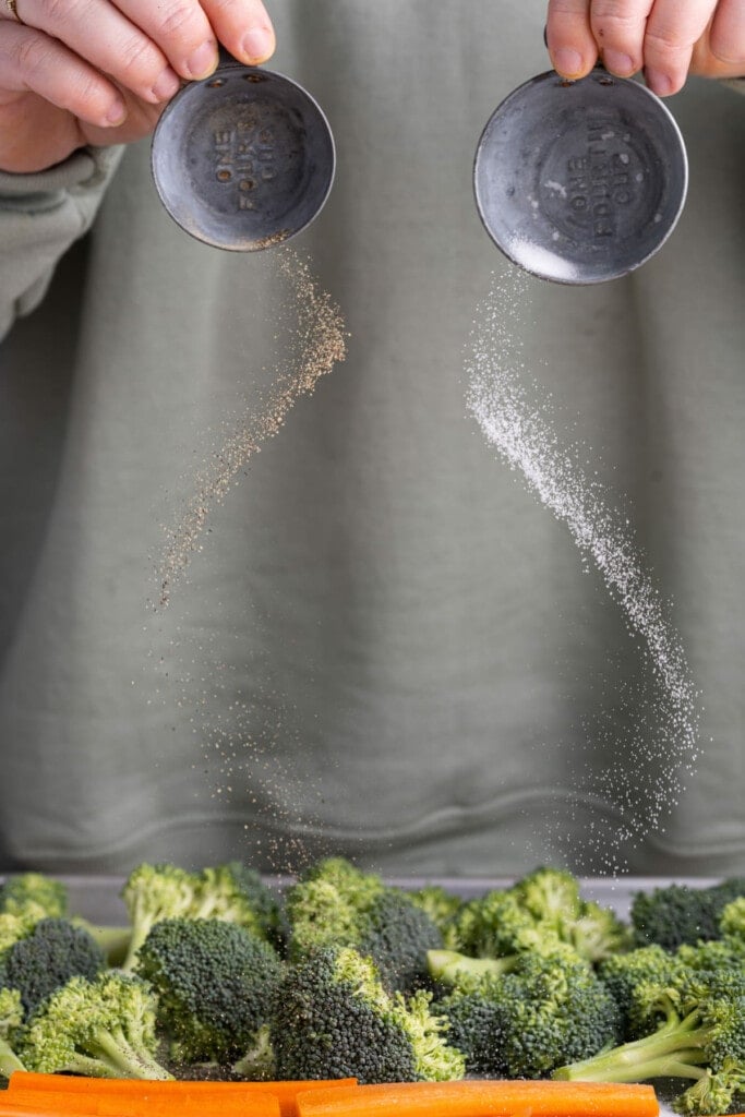 Adding salt and pepper to prepped broccoli and carrots on a baking sheet.