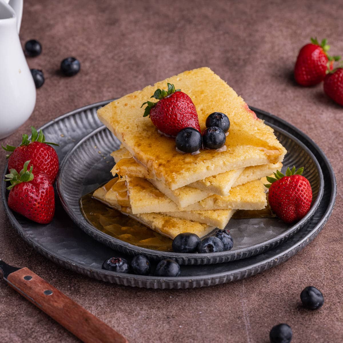 Sheet pan pancakes stacked on a plate with fresh fruit and maple syrup