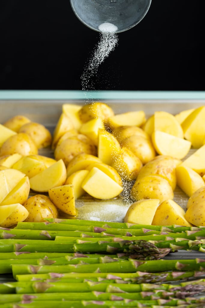 Adding salt to asparagus and potatoes on a sheet pan.
