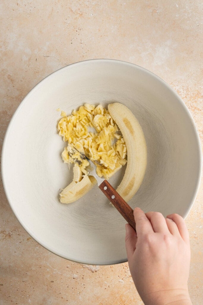 Using a fork to mash ripened banana in a large bowl.