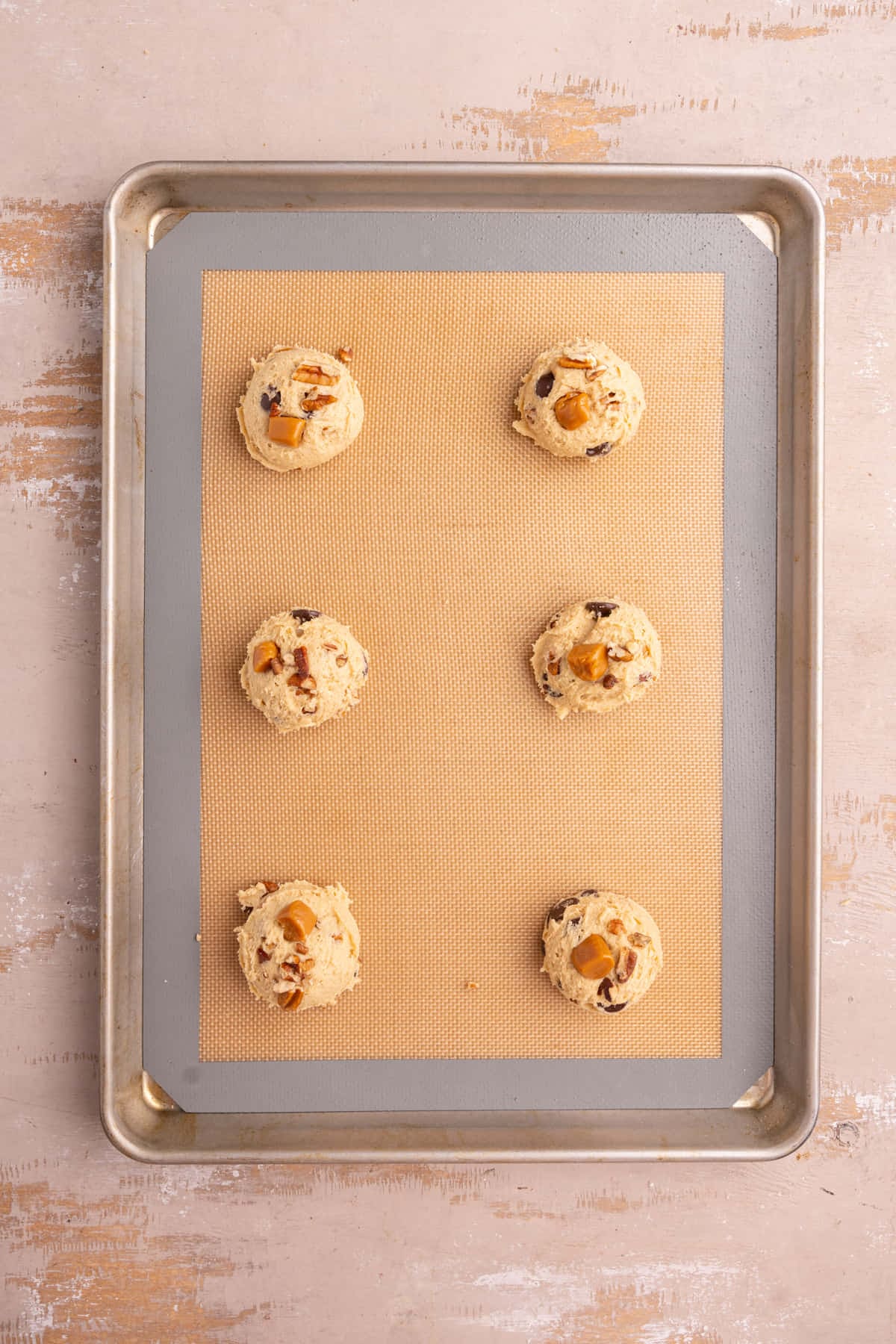 Cookie dough with caramel and pecans and chocolate chips shaped and lined up on a baking sheet