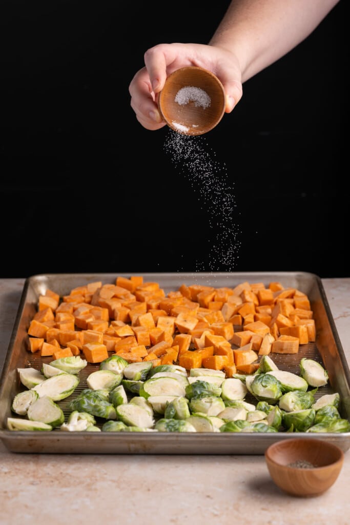 Sprinkling salt on top of brussels sprouts and sweet potatoes on a baking tray.