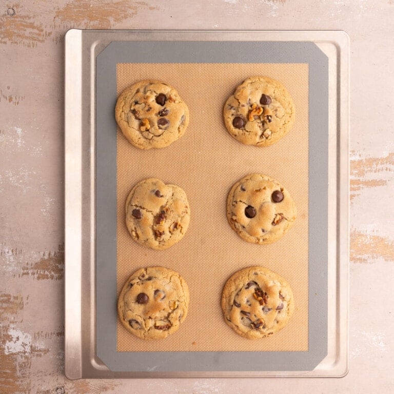 Cookie sheet with a reusable baking mat and six freshly baked chocolate chip walnut cookies.