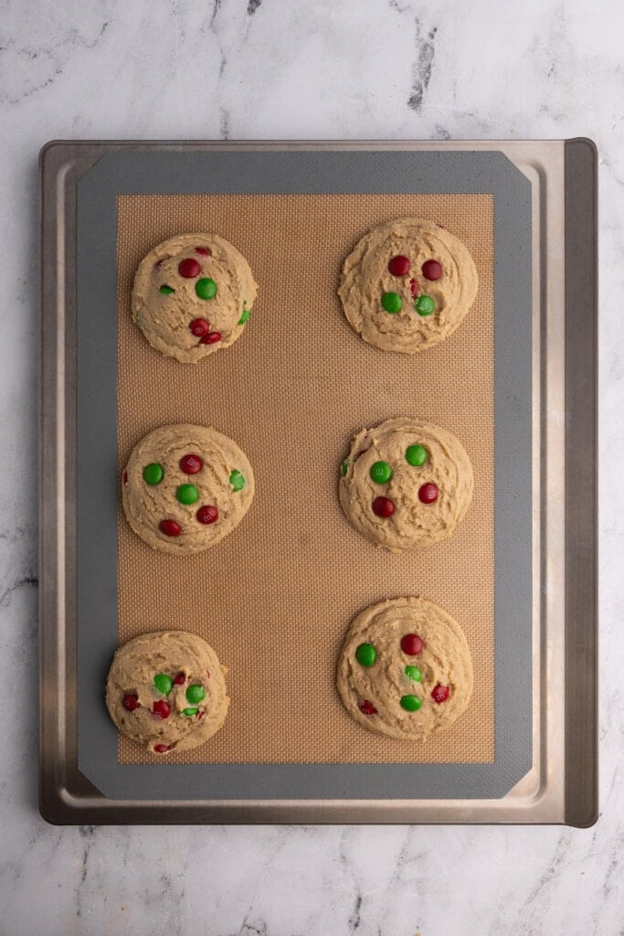Six M&M Christmas cookies lined up on a cookie sheet.