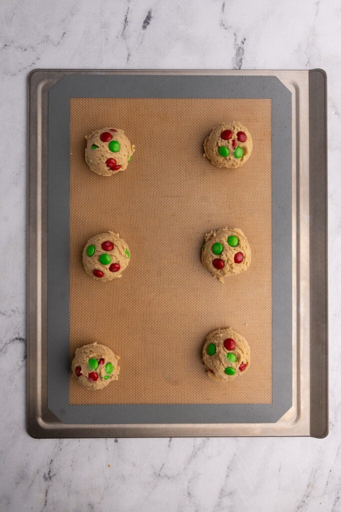 Cookie dough lined up on a lined baking sheet with green and red M&M's pressed into top.