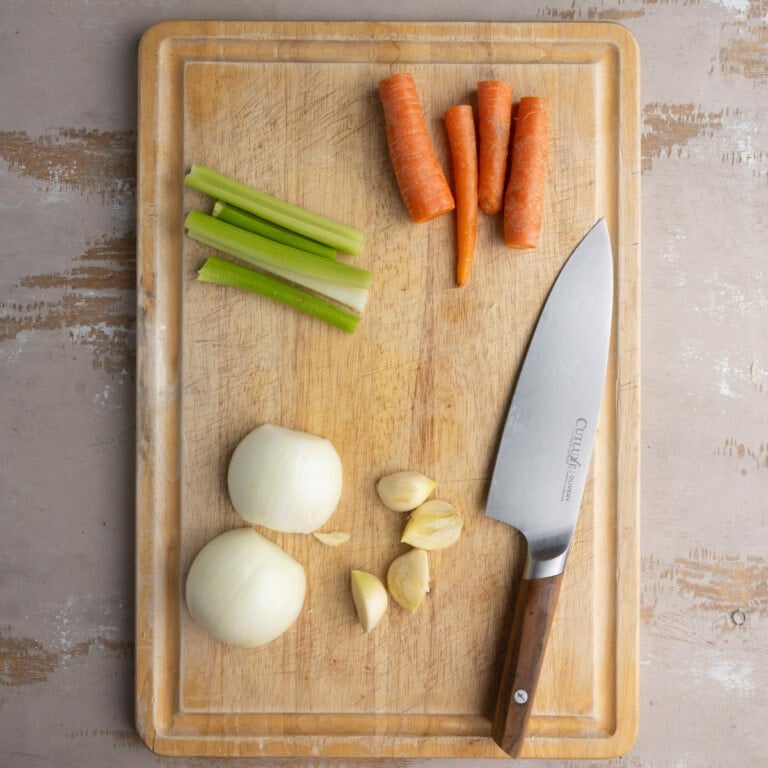 Cleaned and halved carrots and celery and peeled onion and garlic on a chopping board.