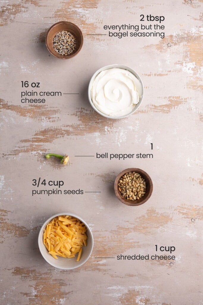 Every ingredient needed to make a Cheese Pumpkin including cream cheese, cheddar cheese, everything but the bagel seasoning, pumpkin seeds, and a bell pepper stem.