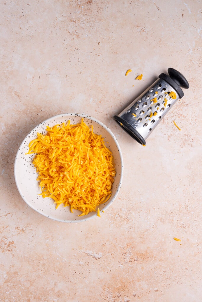 Shredded cheddar cheese in a bowl next to a cheese grater.