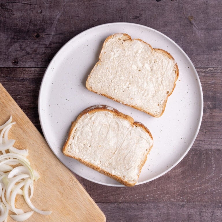 Two slices of bread with even layer of butter and chopping board with sliced onions.