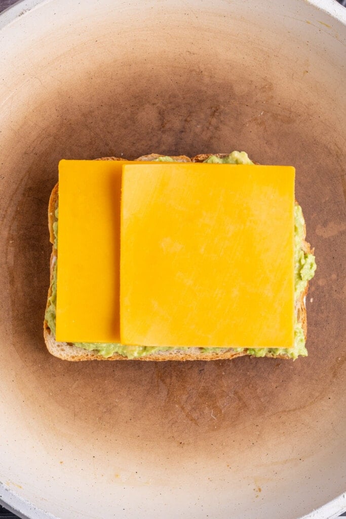 Cheddar cheese slices stacked on top of toasting bread with smashed avocado. 