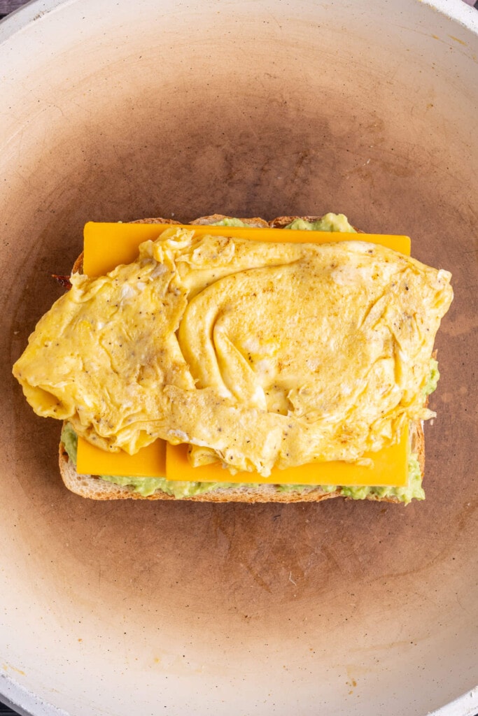 Adding scrambled egg on top of cheddar cheese on smashed avocado spread on toasting bread. 