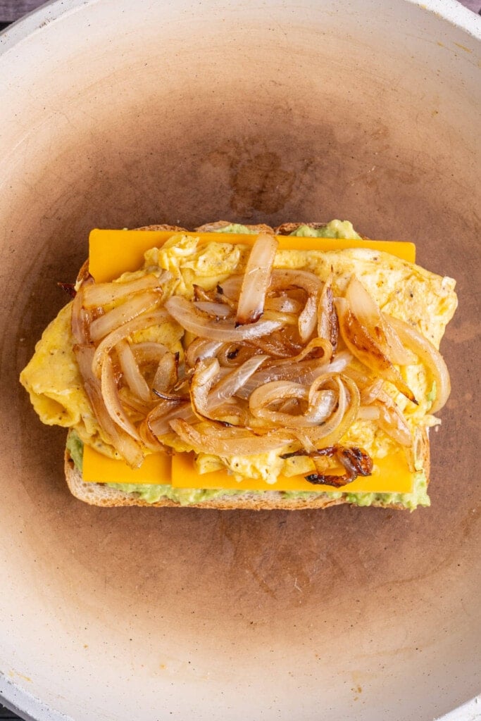 Bread frying in a pan stacked with smashed avocado, cheddar cheese, scrambled eggs, and caramelized onions. 