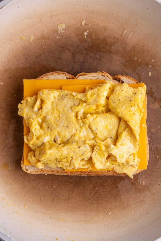 Layering cooked scrambled eggs on top of cheese to assembled breakfast sandwich in frying pan. 