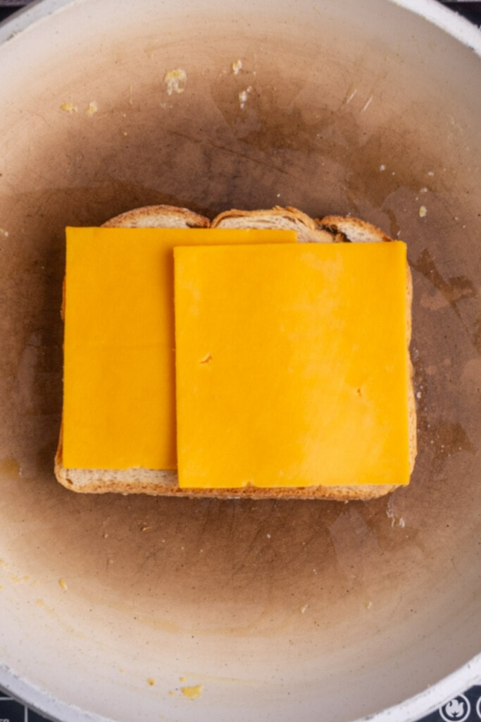 Slices of cheddar cheese stacked on top of buttered bread toasting in a frying pan. 