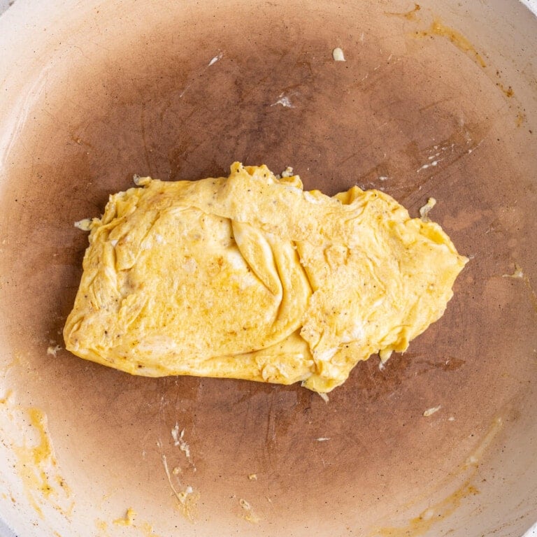Scrambled eggs in the center of a pan, cooked and folded to be roughly the same size of sandwich bread.