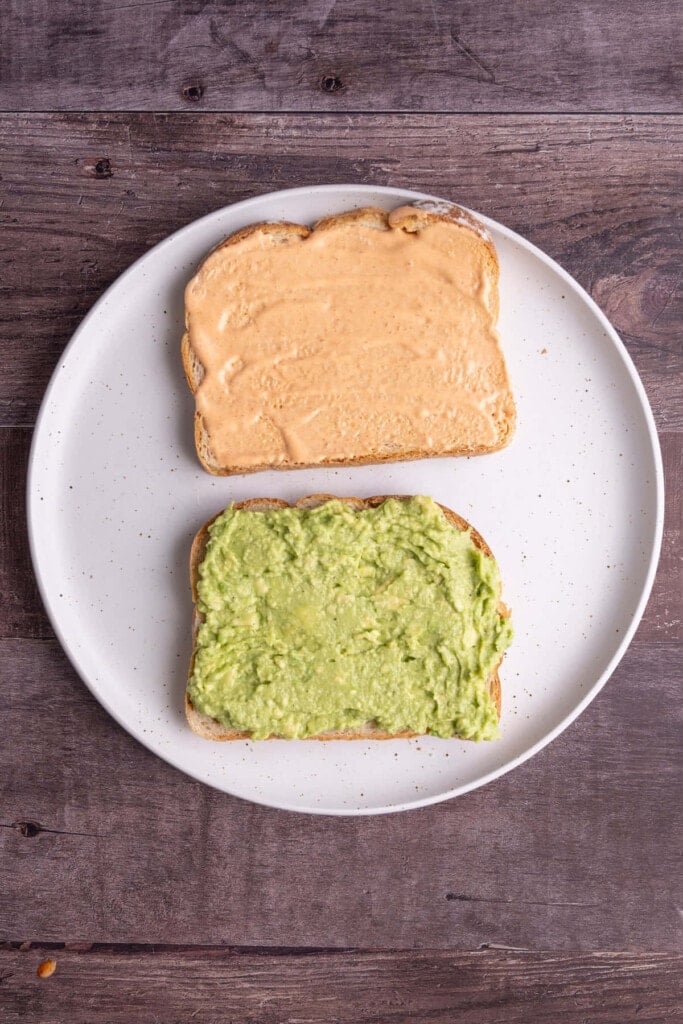 One slice of bread with butter on one side and spicy mayonnaise on the other, and a second slice of bread with butter on one side and smashed avocado on the other.