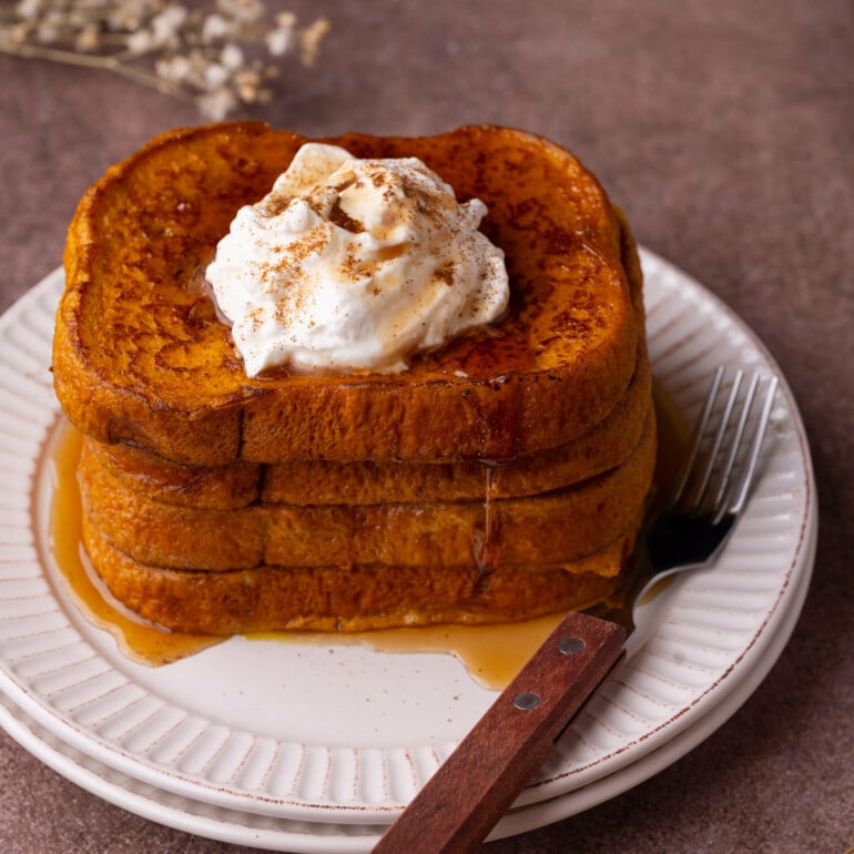 Four slices of Pumpkin French Toast stacked on top of each other with whipped cream and maple syrup.
