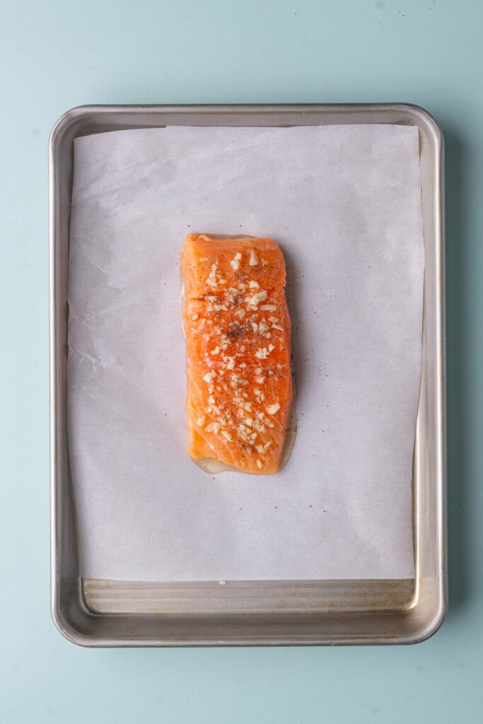 Skinless salmon on a baking sheet lined with parchment paper topped with minced garlic, olive oil, salt, and pepper. 