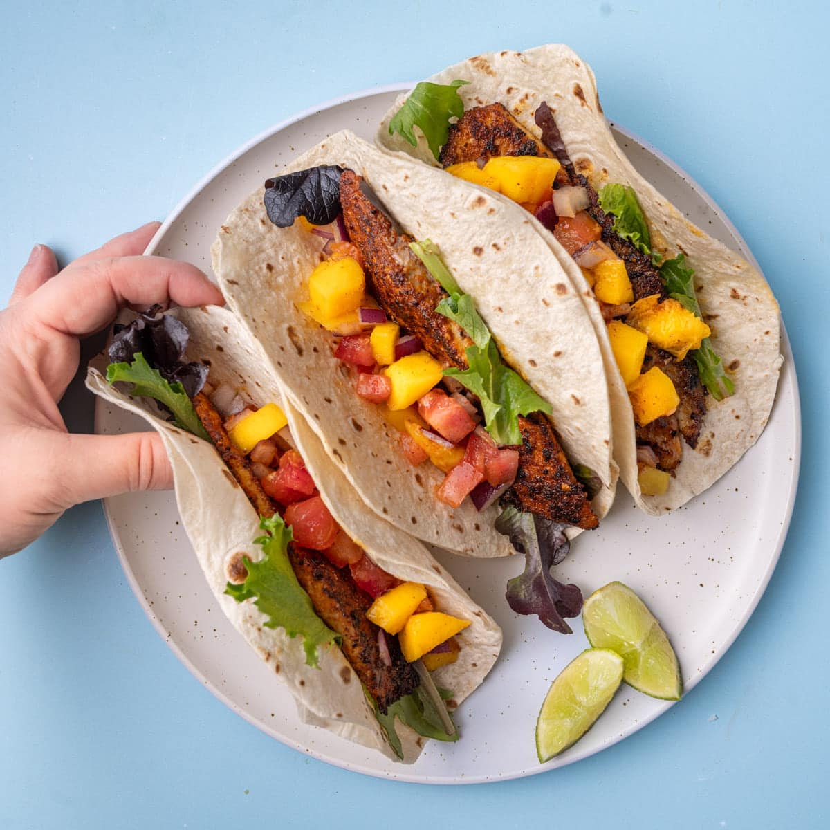 Three fish tacos with mango salsa on a plate, with hand reaching in to grab one.