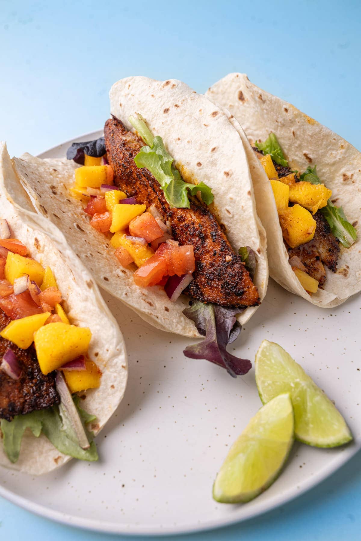 Three mango salsa fish tacos on a large white plate served with lime wedges. 