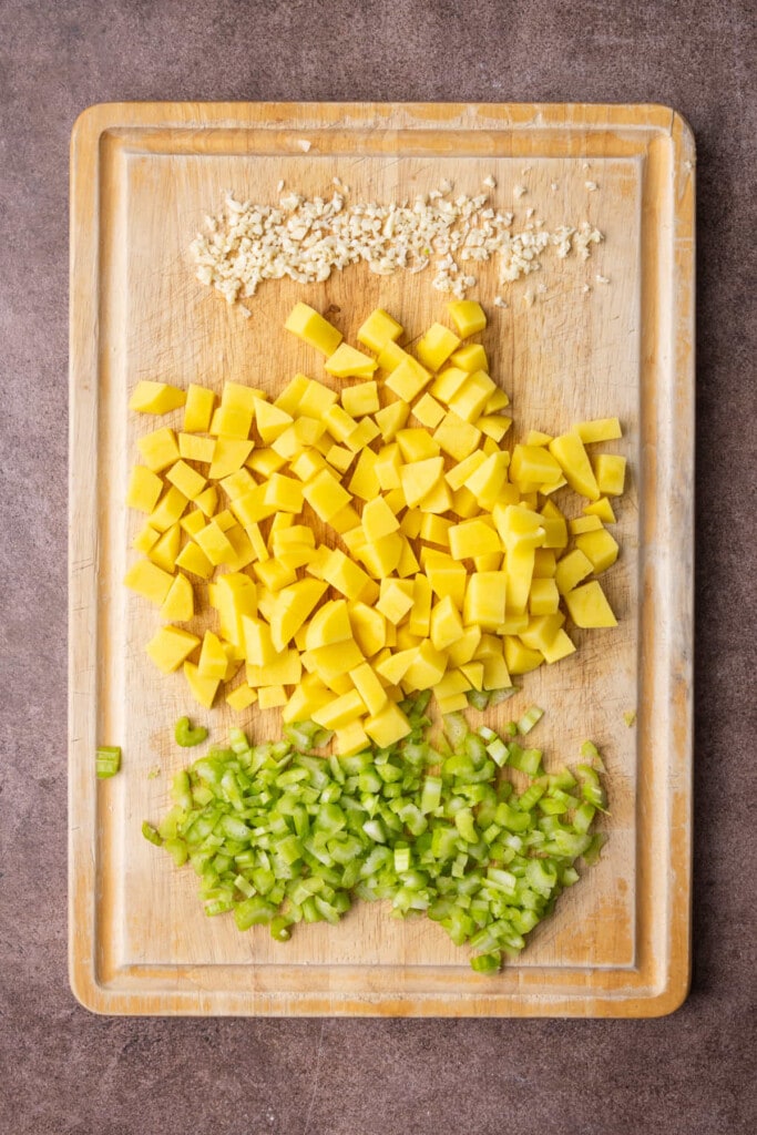 Peeled and minced garlic, peeled and chopped potatoes, and diced celery on a cutting board. 