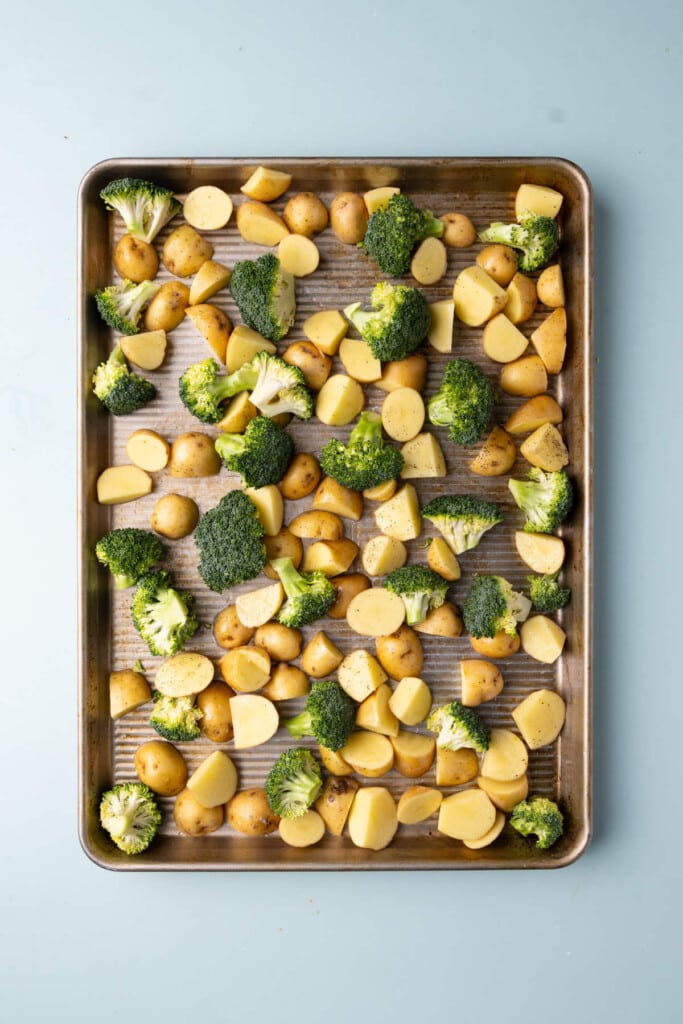 Prepped broccoli and potatoes coated in olive oil, salt, and pepper on a sheet pan.