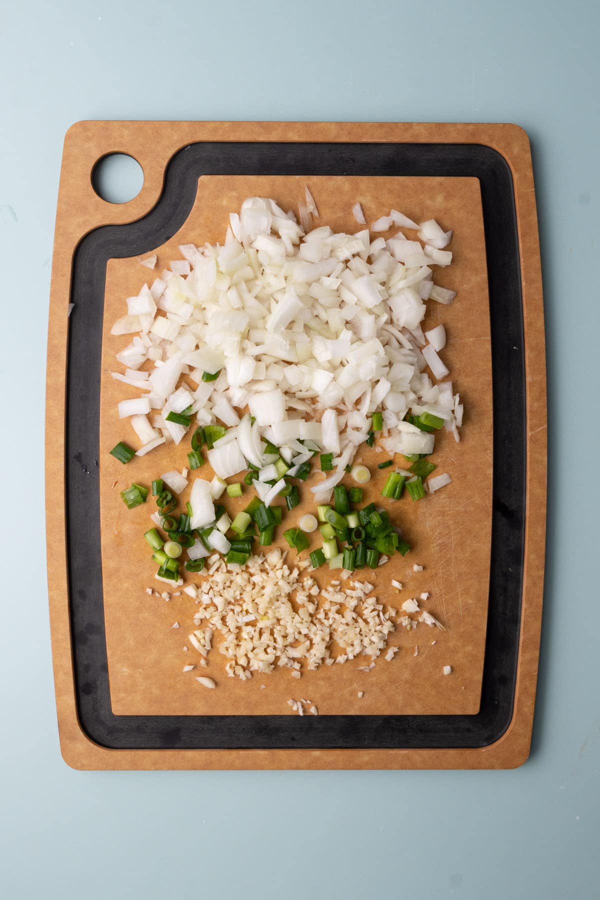 Peeled and diced yellow onion, peeled and minced garlic, and diced scallions on a chopping board. 