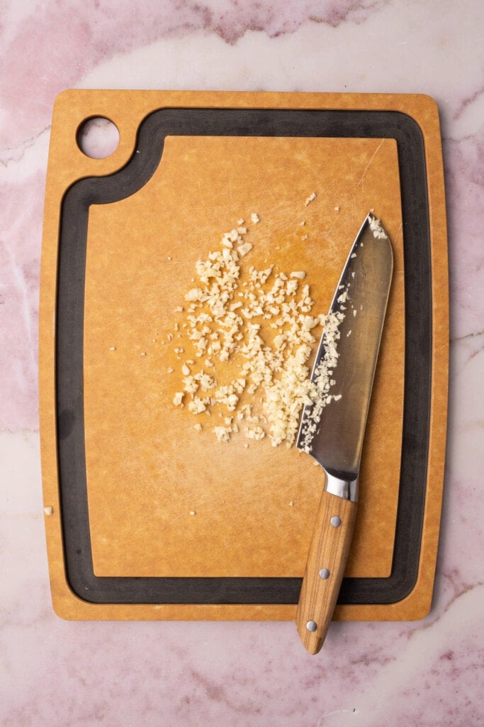 Minced garlic on a chopping board next to a sharp knife. 