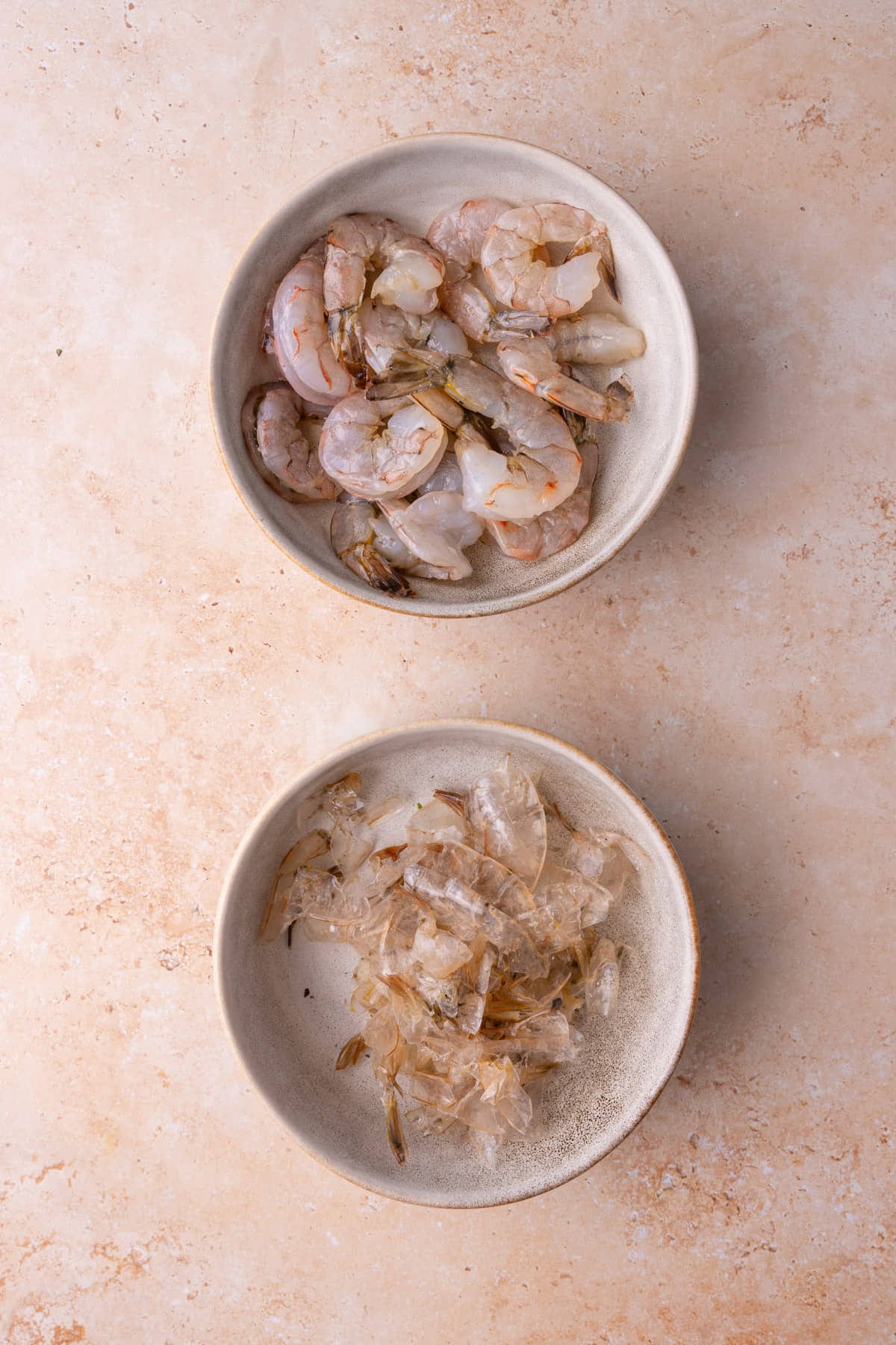 Bowl of peeled, raw, large white shrimp next to a bowl of the discarded peels. 