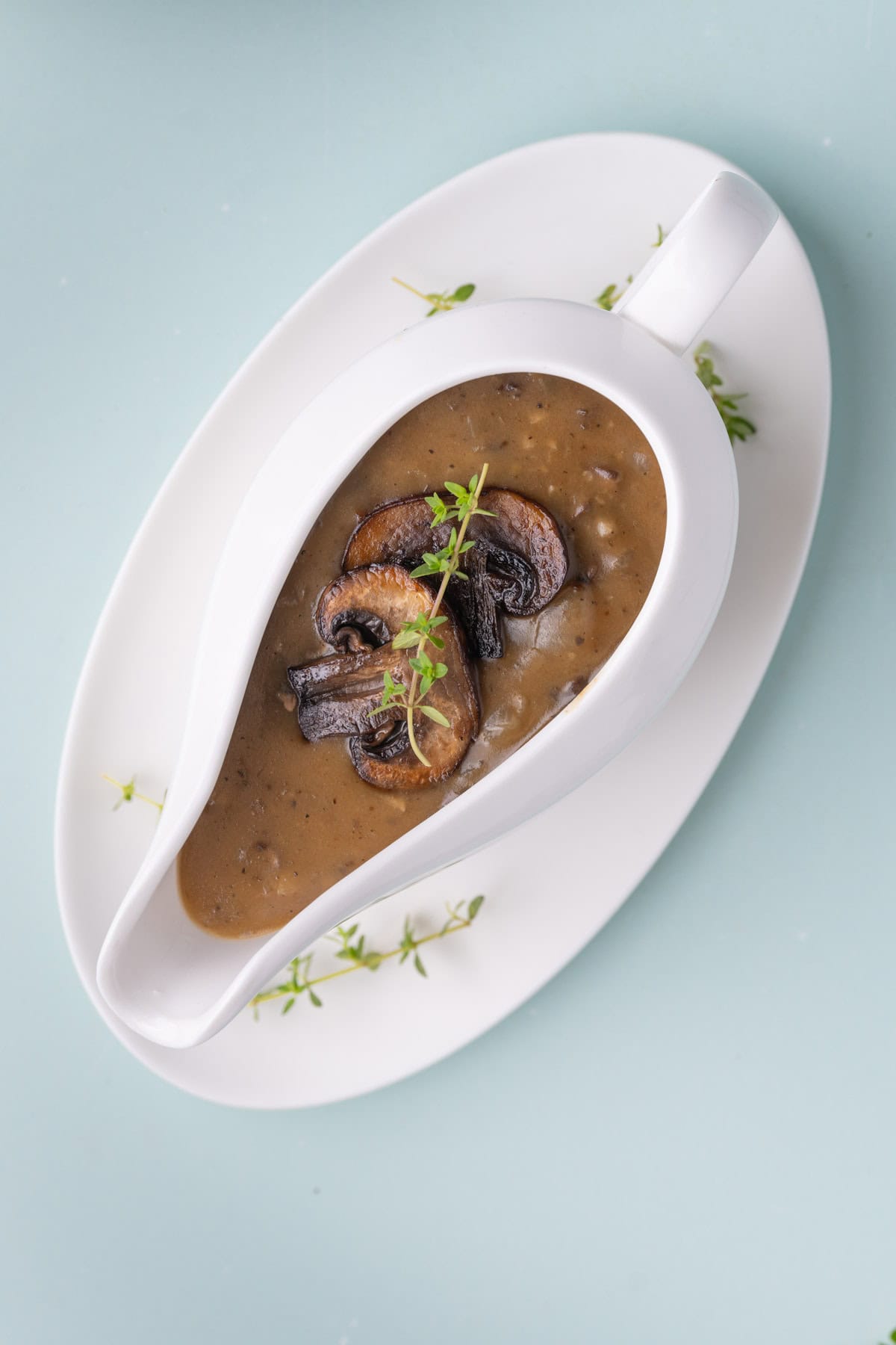 White gravy boat on a dish with mushrooms gravy filled to the top and garnished with fresh thyme.