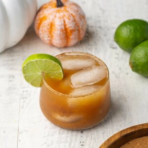 Small glass with an orange spiced margarita in the center of the frame with limes and pumpkins in the background.