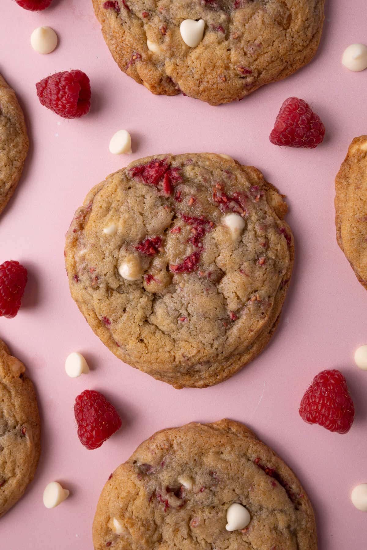 White chocolate raspberry cookies on a pink background surrounded by fresh raspberries and white chocolate chips. 